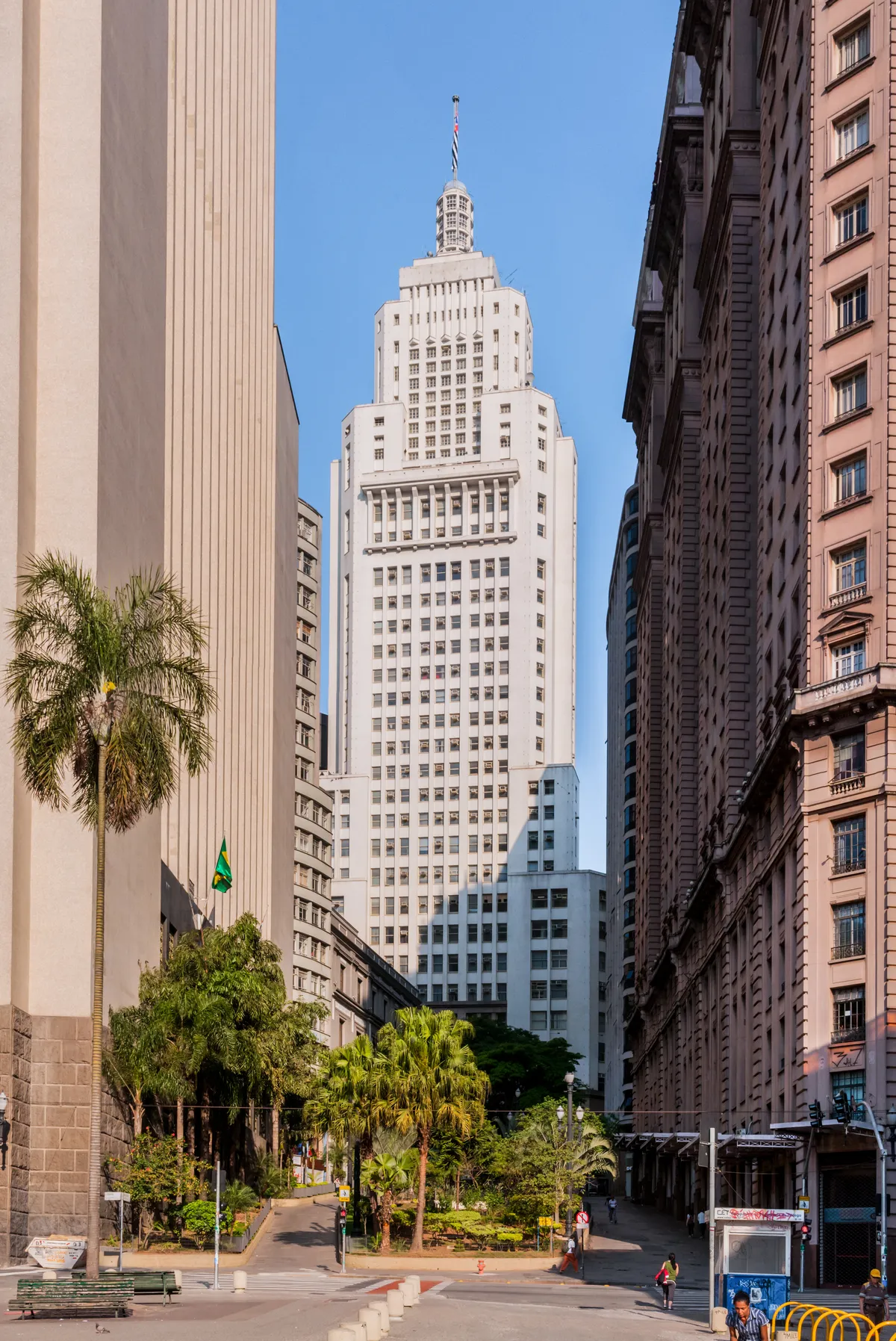 The white Altino Arantes tower rising at the end of a narrow street in São Paulo's banking district, its stepped upper floors and mast catching the sun above lower stone buildings and palm trees