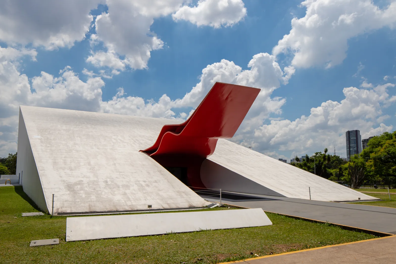 The Ibirapuera Auditorium in São Paulo, a low white concrete wedge on grass with a folded red steel canopy curling out over its entrance under a bright cloudy sky