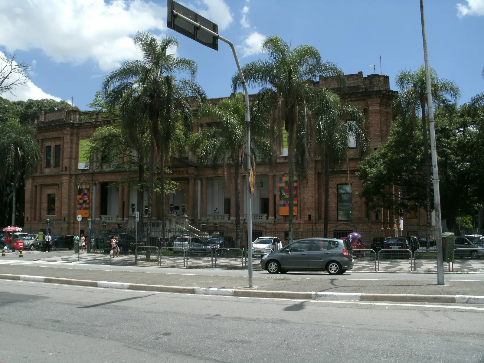 The long bare-brick facade of the Pinacoteca art museum in São Paulo seen from Avenida Tiradentes, with tall palms along the pavement in front of it and traffic on the avenue between