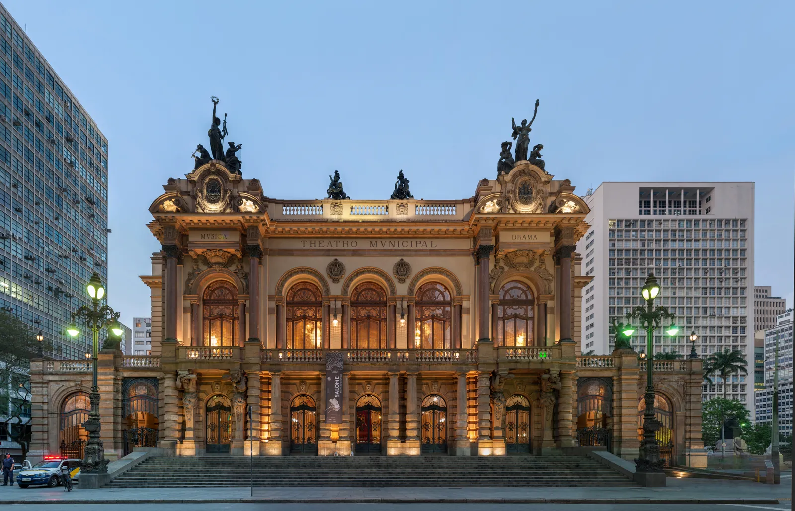 The lit facade of the Theatro Municipal in São Paulo at dusk, an ornate stone opera house with arched windows, columns and bronze figures along the roofline, framed by modern office blocks on either side
