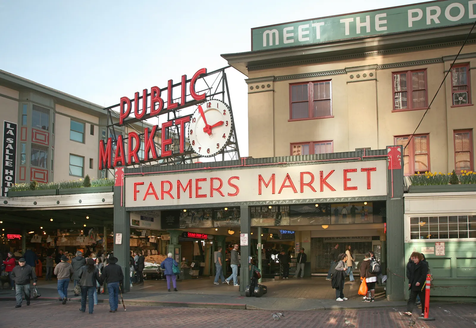 The neon Public Market Center sign and brass clock over the main entrance to Pike Place Market in Seattle, with the Farmers Market arcade and shoppers below