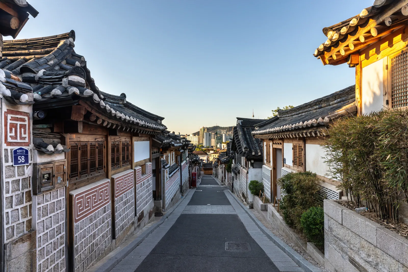 A lane in Bukchon Hanok Village in Seoul lined with traditional hanok houses, grey stone walls, wooden gates and curved tiled roofs, running downhill toward the city