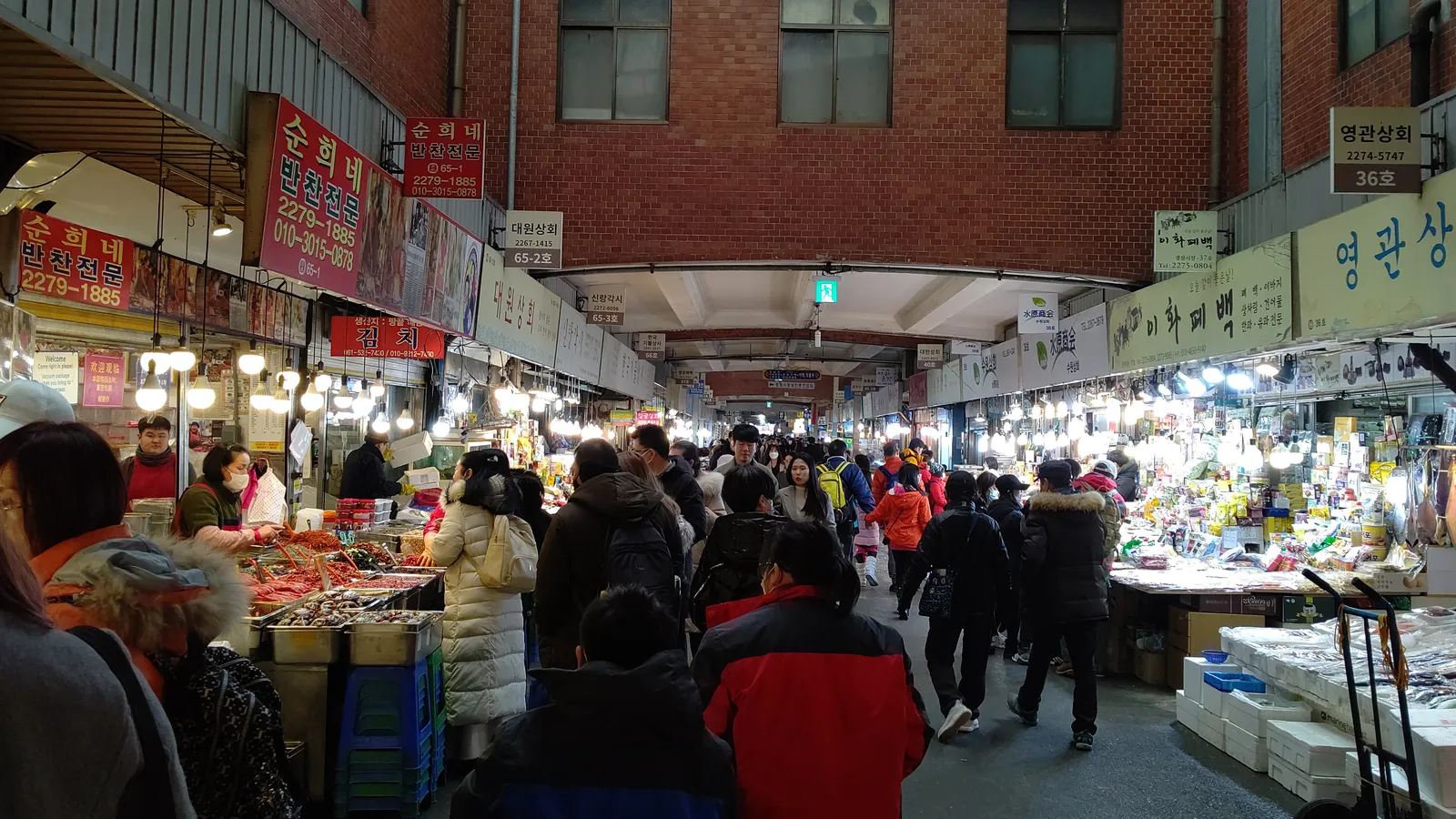 The covered main aisle of Gwangjang Market in Seoul, crowded with shoppers between brightly lit food and produce stalls under a high roof