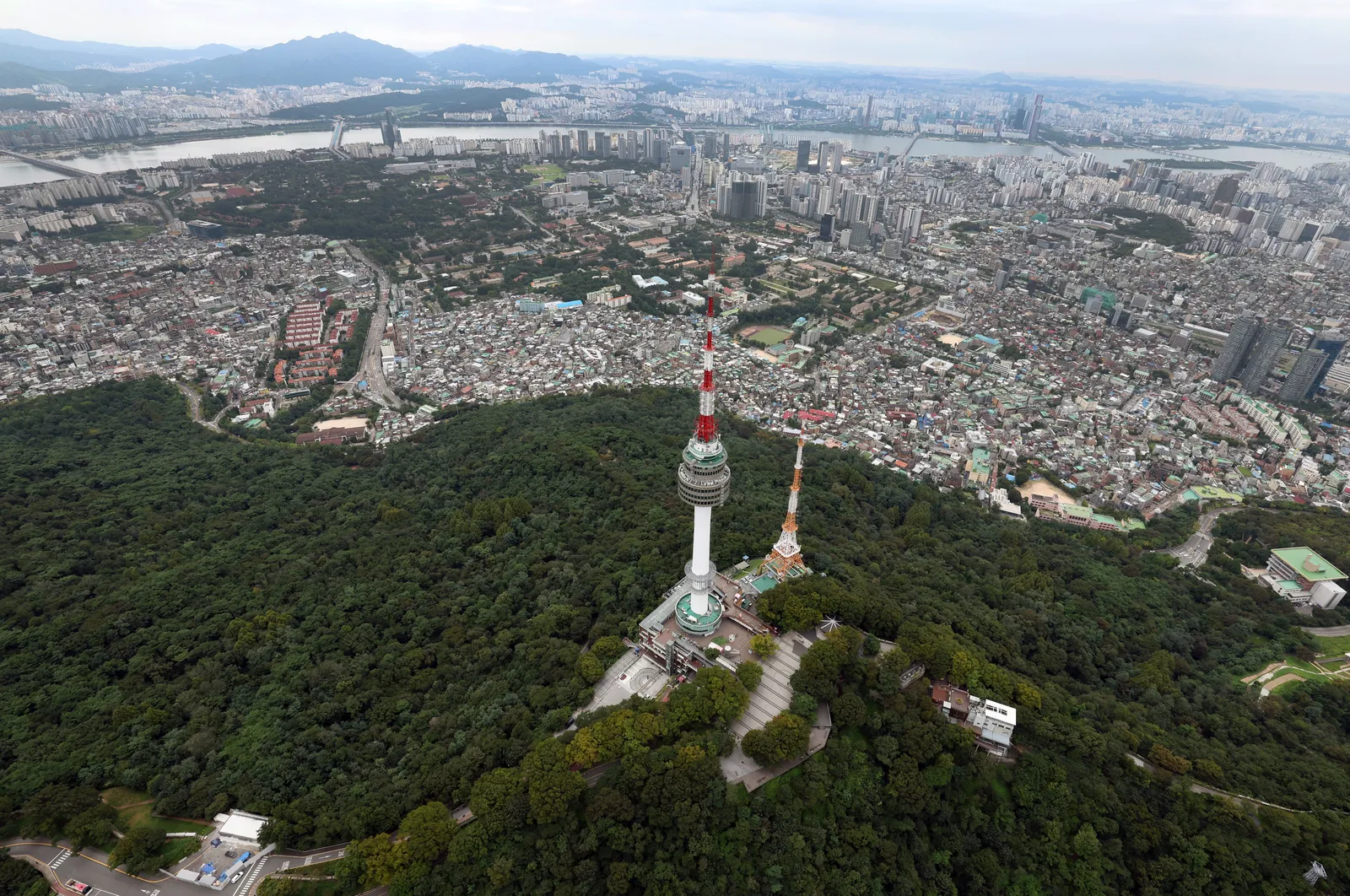 Aerial view of N Seoul Tower on the wooded summit of Namsan, with dense city blocks spreading out around the hill and the Han River beyond