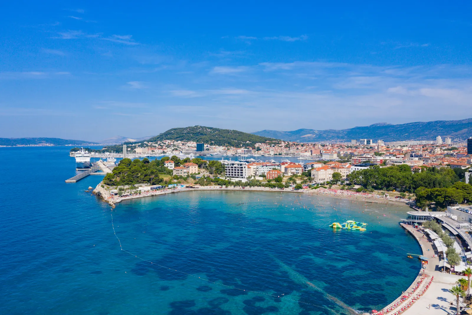 Bačvice bay in Split from the air, a curve of pale sand and shallow turquoise water with swimmers in it, the city and its ferry berths on the headland behind