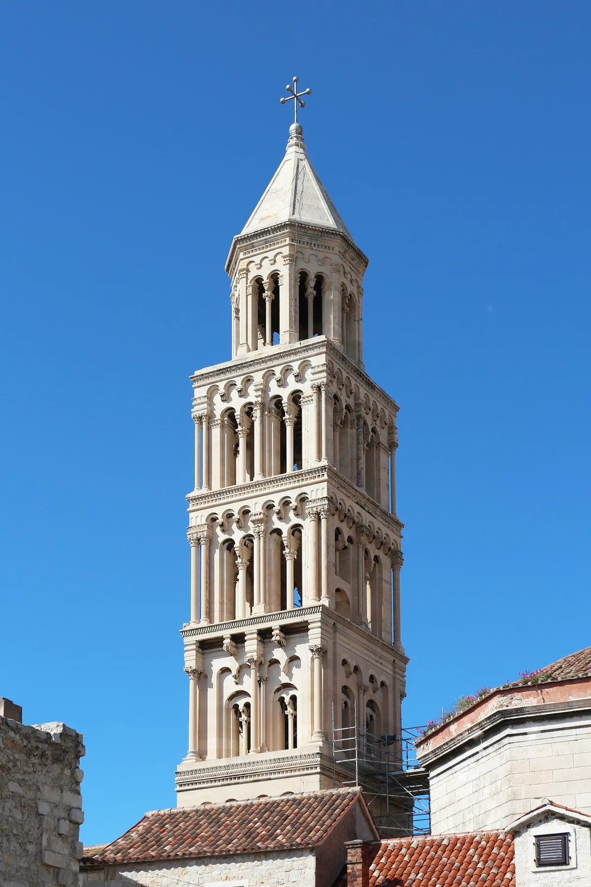 The bell tower of Split's cathedral against a clear blue sky, a tall pale stone tower of stacked arcaded storeys narrowing to a small pyramid spire, with terracotta roofs at its base