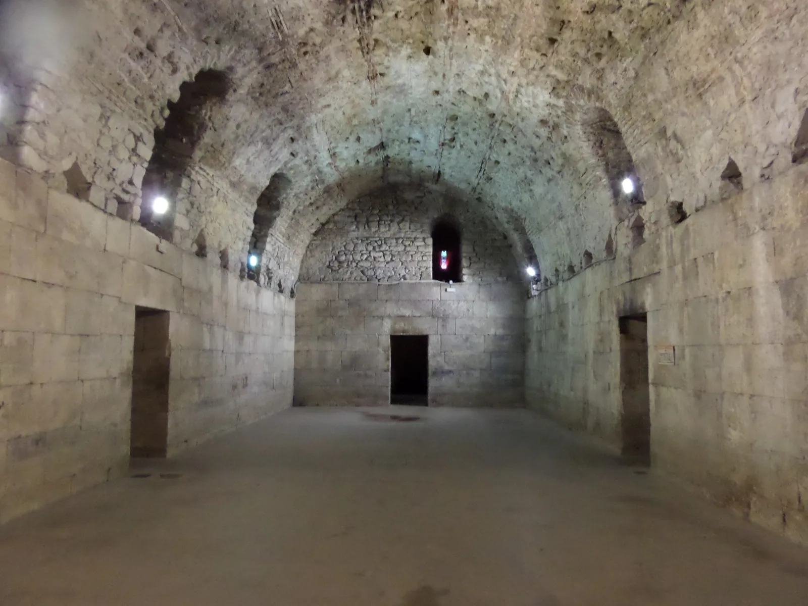 A barrel-vaulted stone hall in the cellars beneath Diocletian's Palace in Split, bare limestone walls and a curved ceiling lit by small lamps, with an empty floor running to a doorway at the far end