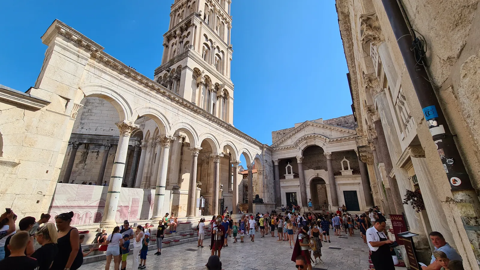 The Peristyle courtyard inside Diocletian's Palace in Split, rows of tall stone columns and arches on either side of a sunken square of flagstones, with the cathedral bell tower rising behind them and people standing on the steps