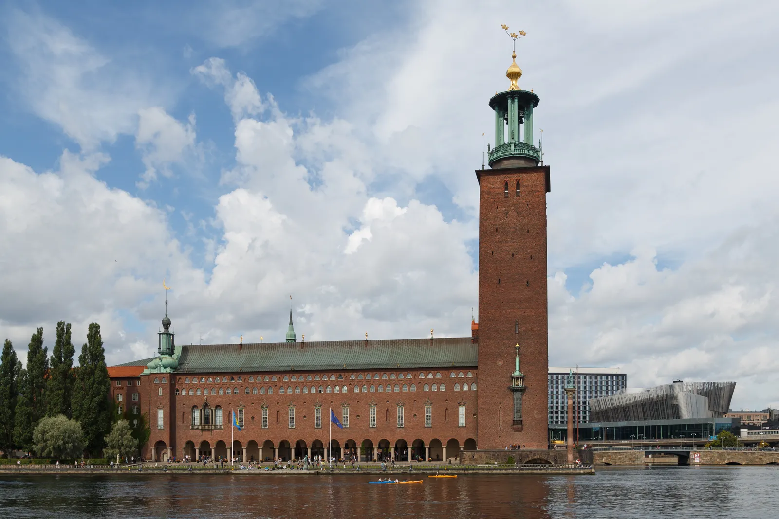 Stockholm City Hall seen across the water, a long dark red brick building with an arcaded waterfront front and a square tower topped by a green copper lantern and three golden crowns