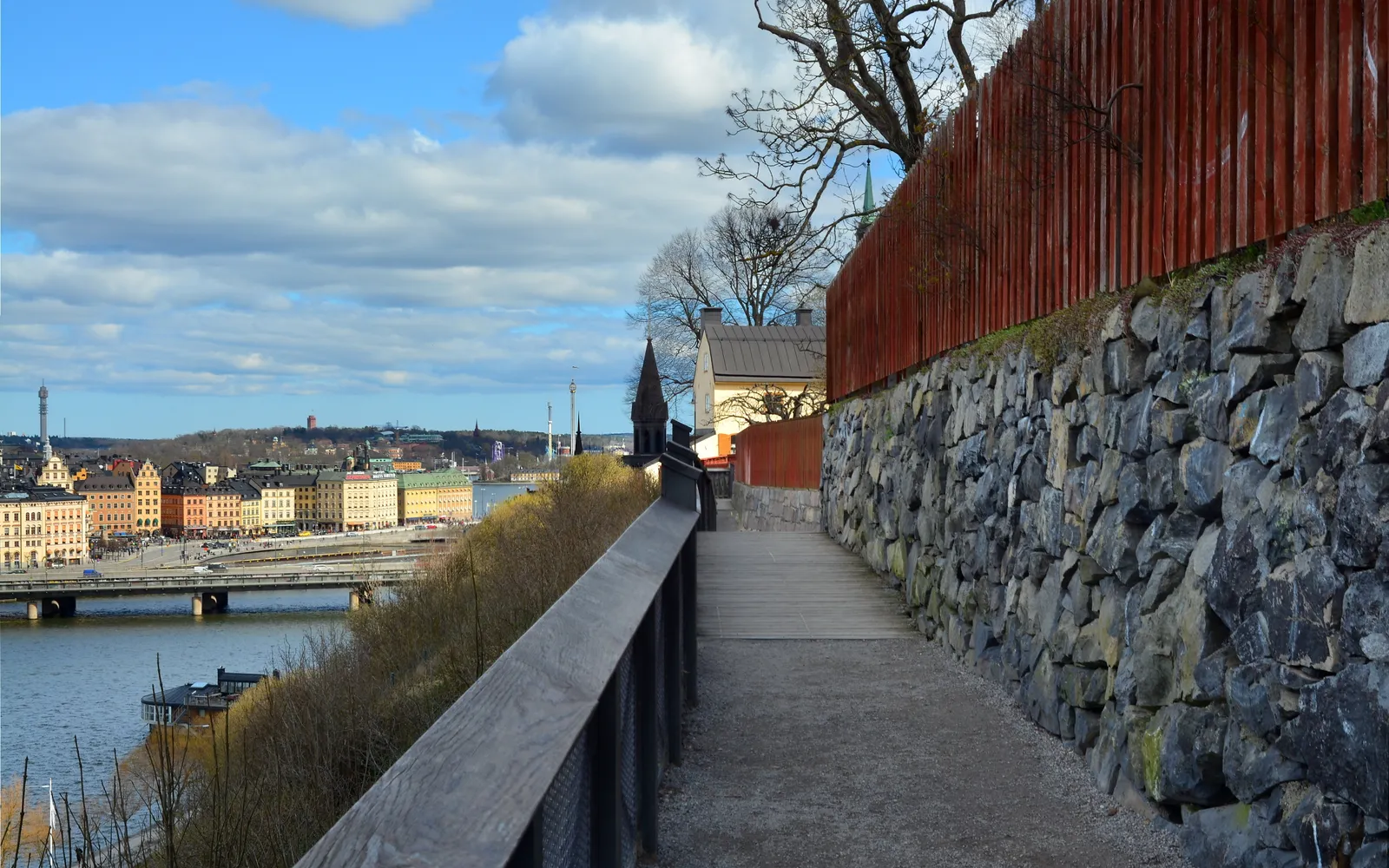 The Monteliusvägen walkway in Stockholm, a narrow wooden path with a railing running between a red timber fence and a stone wall along a clifftop, with the water and the rooftops of the city visible below to the left