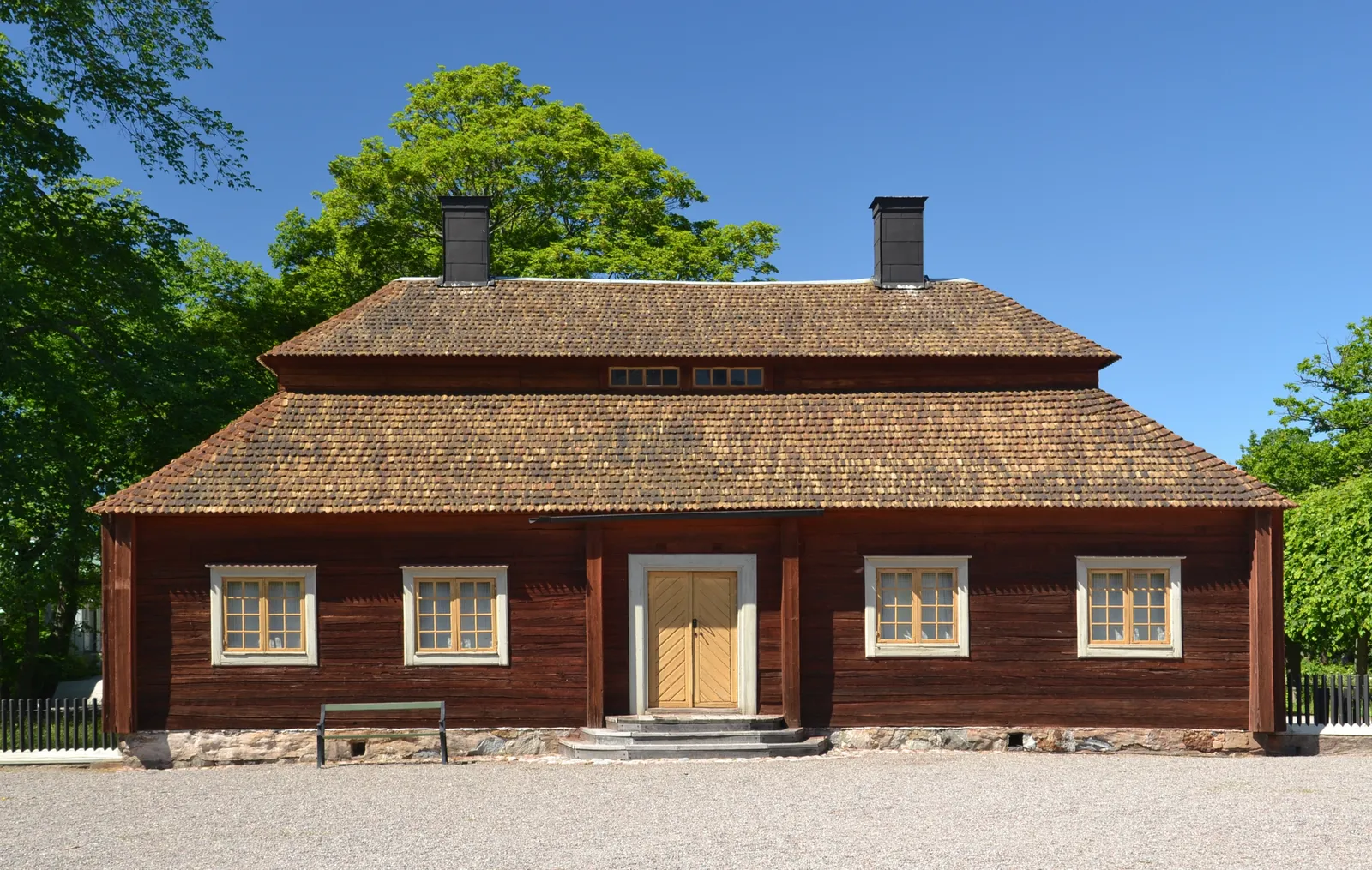 A long red timber manor house with a wooden shingle roof and white-framed windows at Skansen in Stockholm, standing on gravel under trees