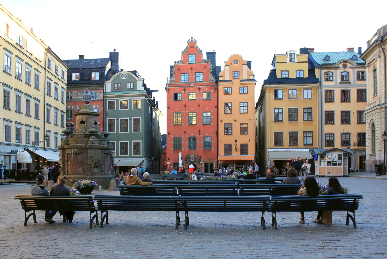 Stortorget square in Stockholm's old town, tall narrow merchant houses in red, ochre, yellow and cream standing shoulder to shoulder behind a stone well, with people sitting on benches on the cobbles