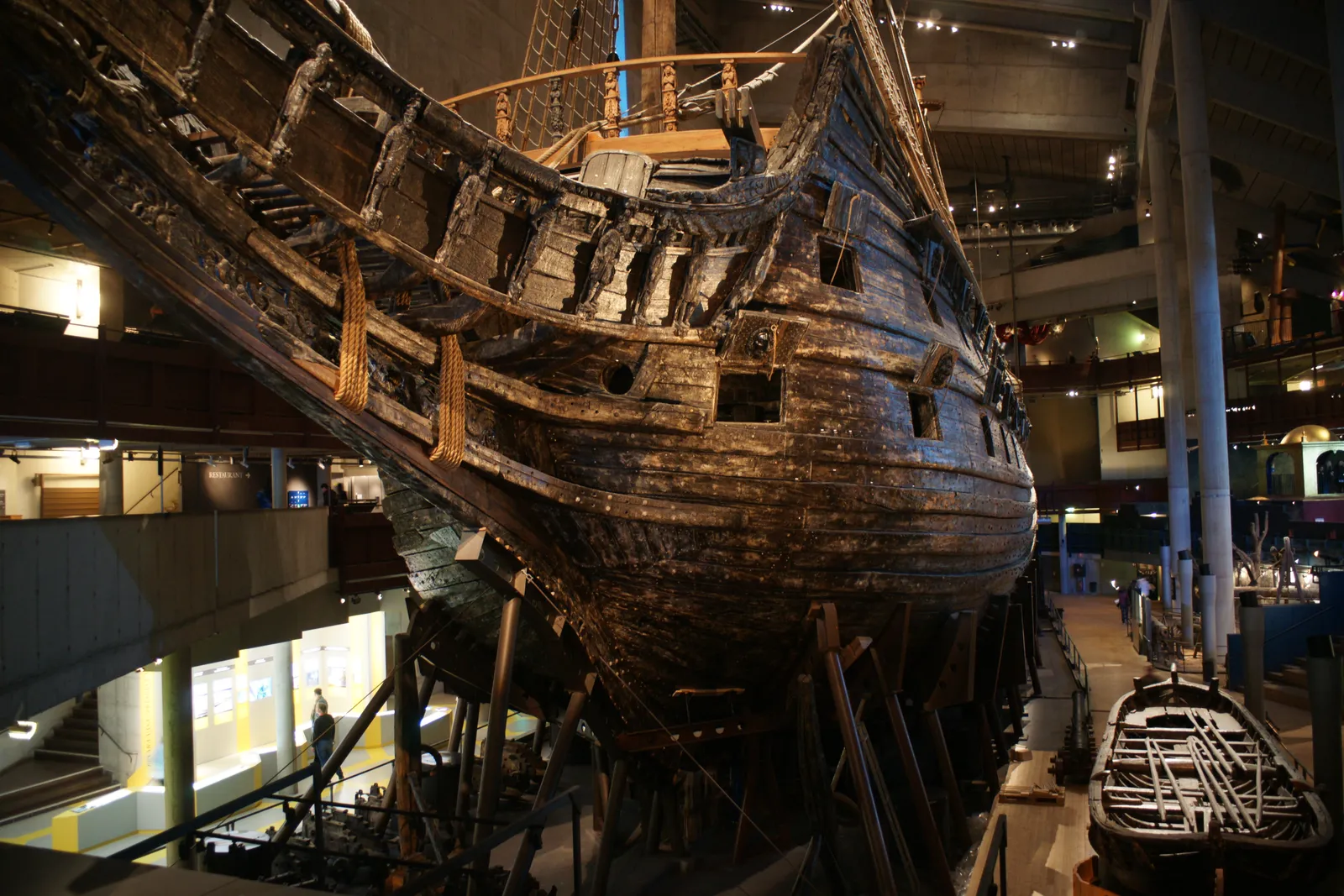 The warship Vasa seen from its bow inside the Vasa Museum in Stockholm, the blackened oak hull rising several storeys above the museum floor with galleries and visitors small alongside it