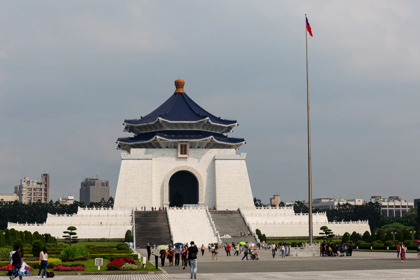 The white marble Chiang Kai-shek Memorial Hall under its octagonal blue roof, with the national flag on a tall pole beside it and visitors crossing the paving in front