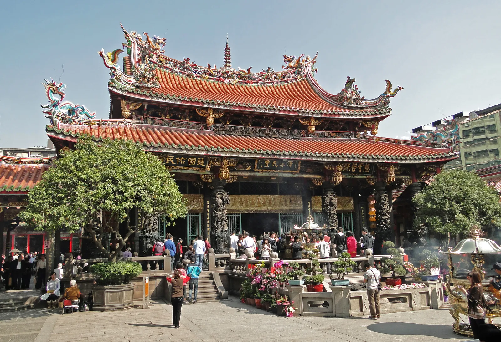 The main hall of Longshan Temple in Taipei, its tiled roofs crowded with dragon and phoenix figures, with worshippers standing at the incense burner in the courtyard