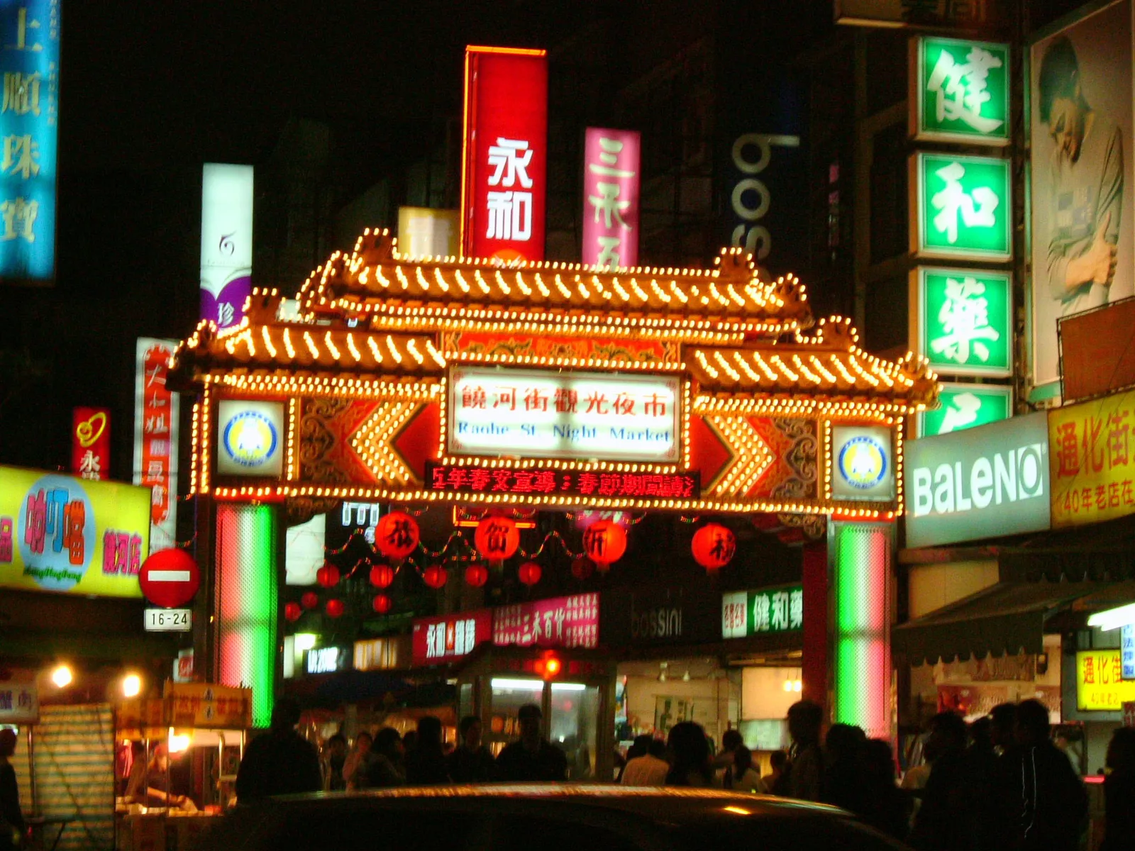 The lit entrance arch of Raohe Street Night Market in Taipei at night, ringed with bulbs and red lanterns, with neon shop signs and a crowd in the street beneath it