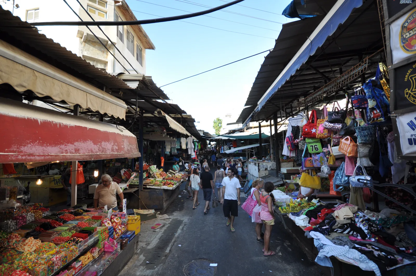 The single aisle of Carmel Market in Tel Aviv, shoppers walking between produce stalls and clothing racks under canvas awnings strung across the lane