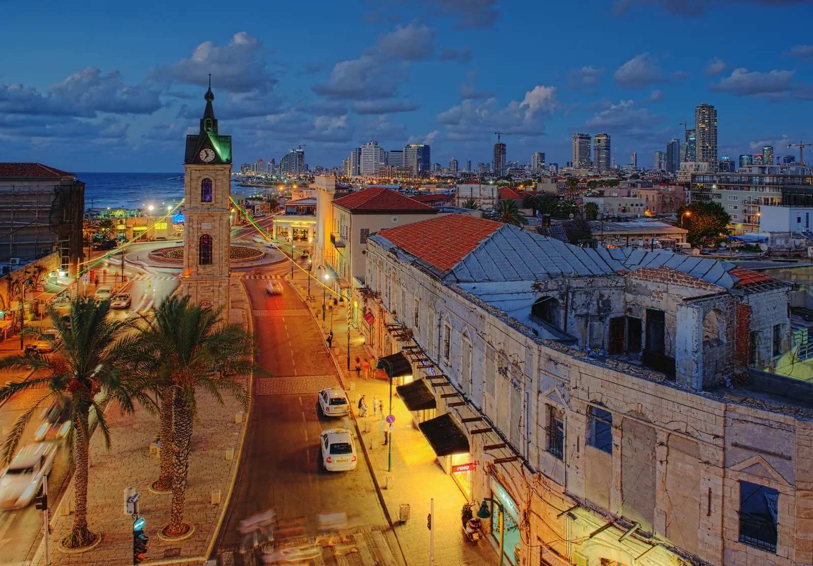 The limestone clock tower at the head of Yefet Street in Jaffa lit up at dusk, with the low arcaded buildings of the old town around it and the towers of central Tel Aviv along the coast behind