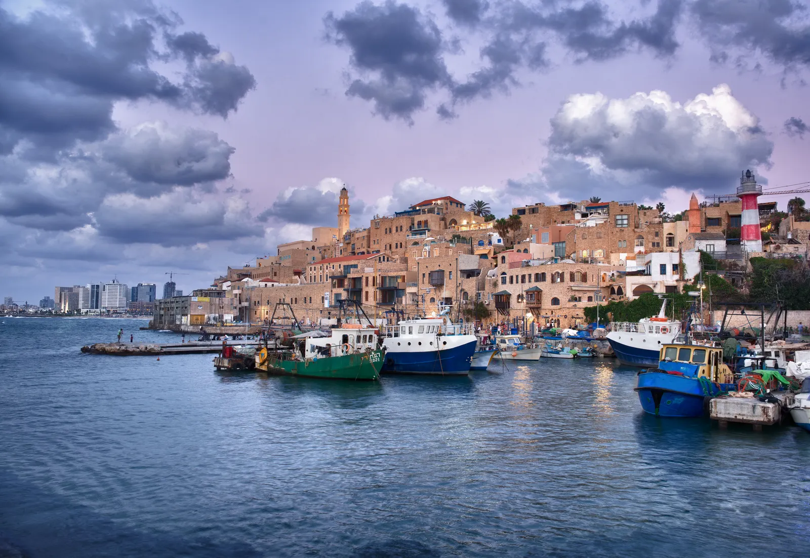 Fishing boats moored in Jaffa's harbour at dusk, with the sandstone houses of the old town stacked up the hill behind them and a red and white lighthouse on the right