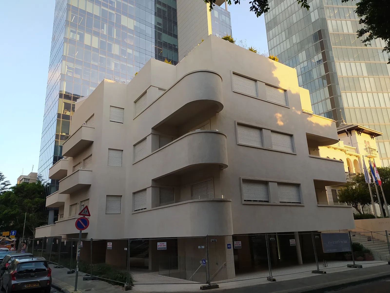 A pale 1930s International Style block on Rothschild Boulevard in Tel Aviv, its corner balconies wrapping the building in smooth curves, with a glass office tower rising behind it