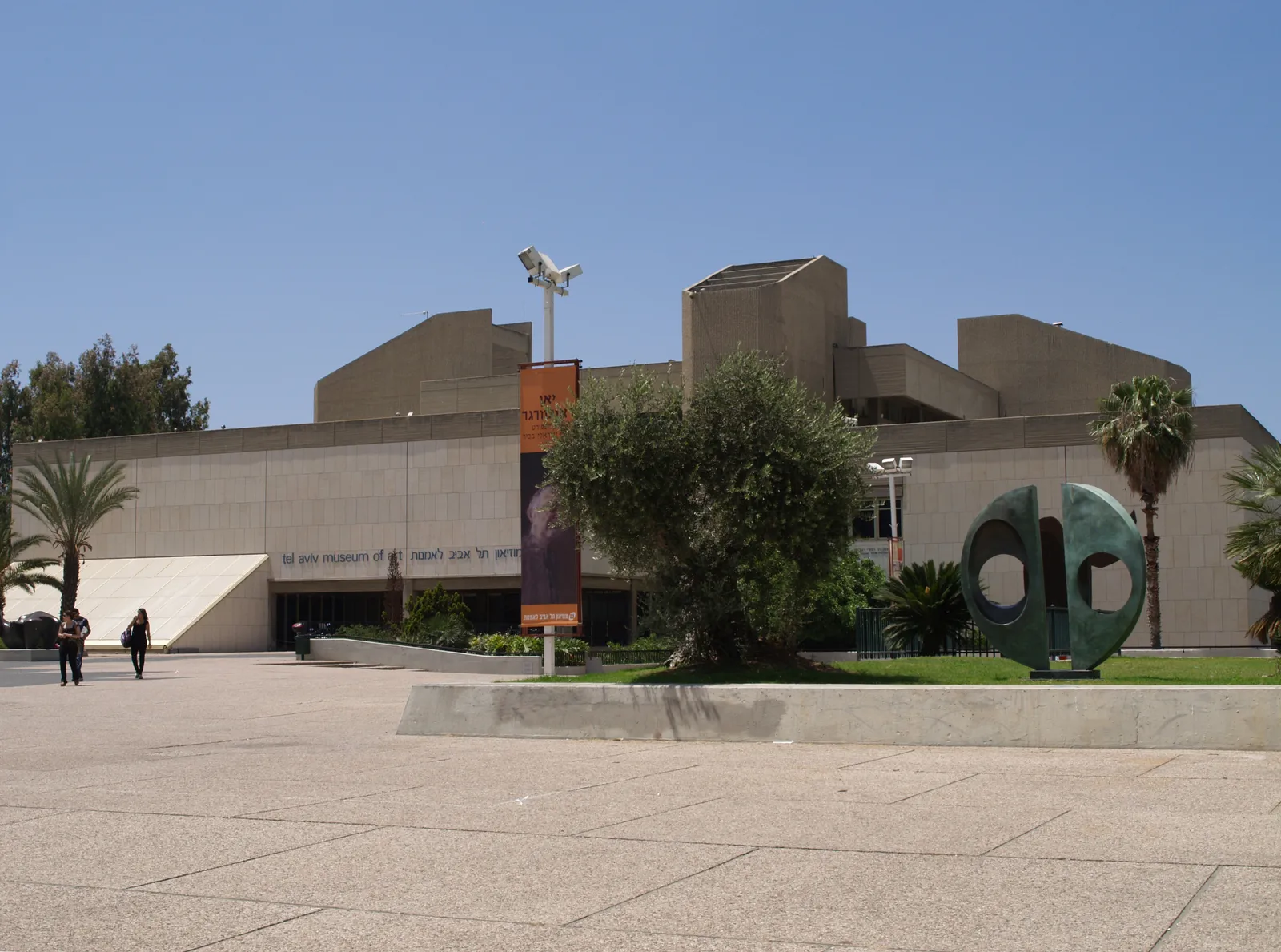 The long sand-coloured concrete frontage of the Tel Aviv Museum of Art seen across its paved forecourt, with olive trees and palms in front of the entrance
