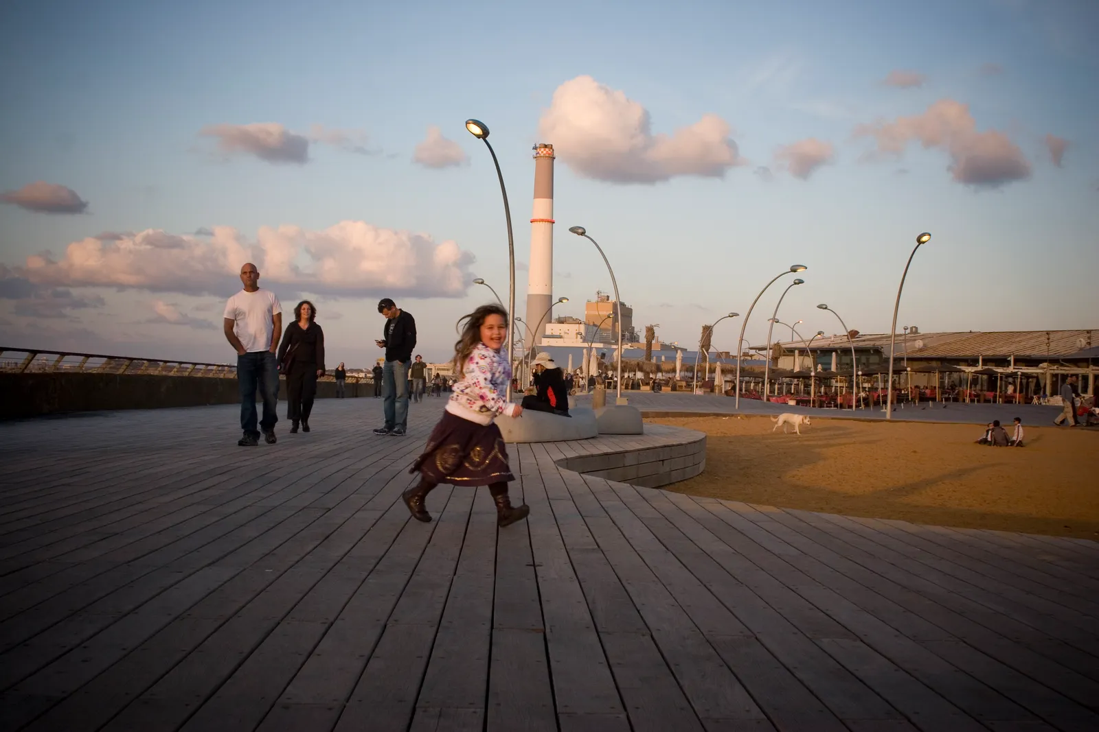 The undulating wooden deck at the old Tel Aviv port in late afternoon light, with people walking across it, curved lamp posts along its edge and a tall chimney on the skyline behind