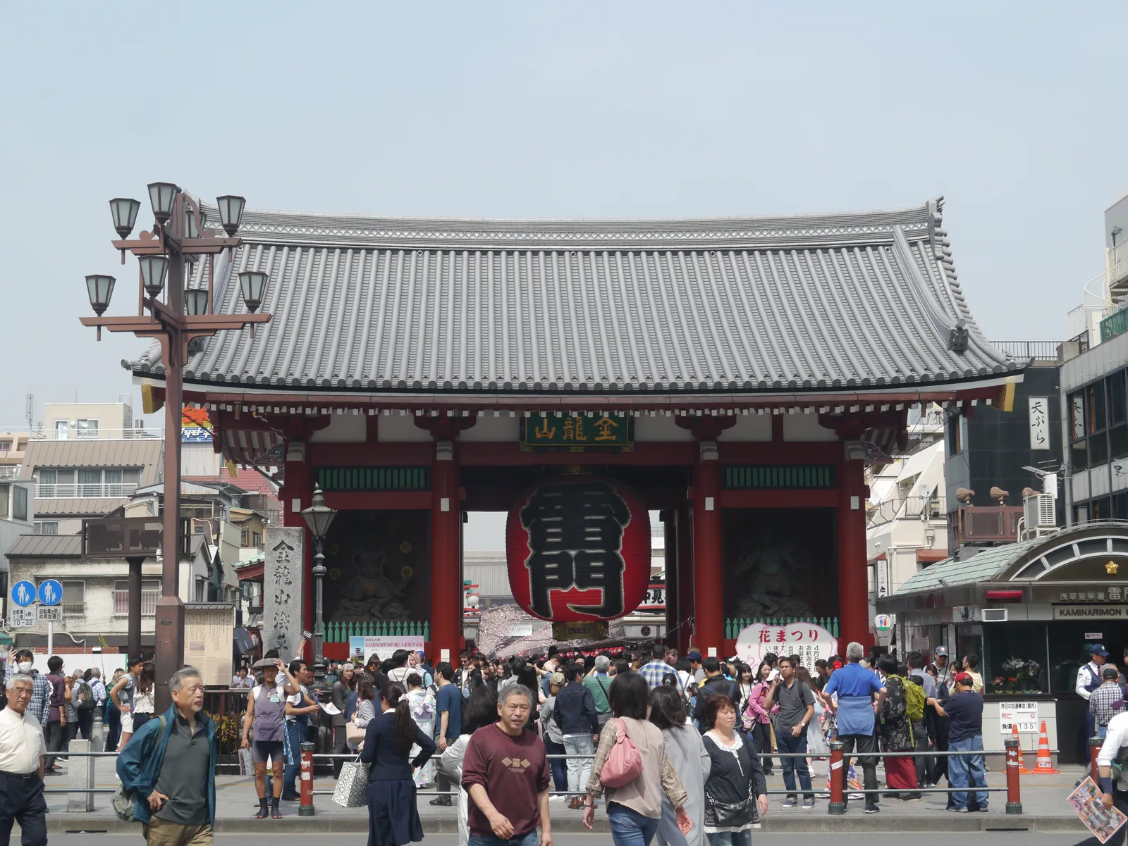 The Kaminarimon gate at Senso-ji in Asakusa, a red timber gate under a grey tiled roof with a large red paper lantern hanging in the central opening and a crowd filling the square in front of it