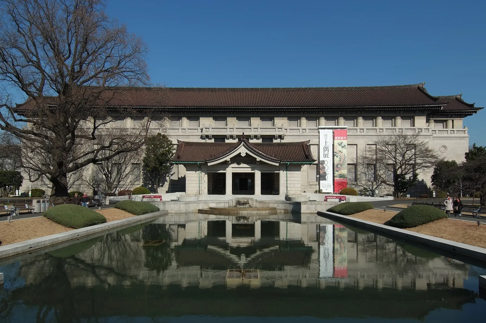 The Honkan building at Tokyo National Museum, a long pale stone gallery under a dark tiled roof with a pillared entrance porch in the middle, reflected in the rectangular pool set in the forecourt