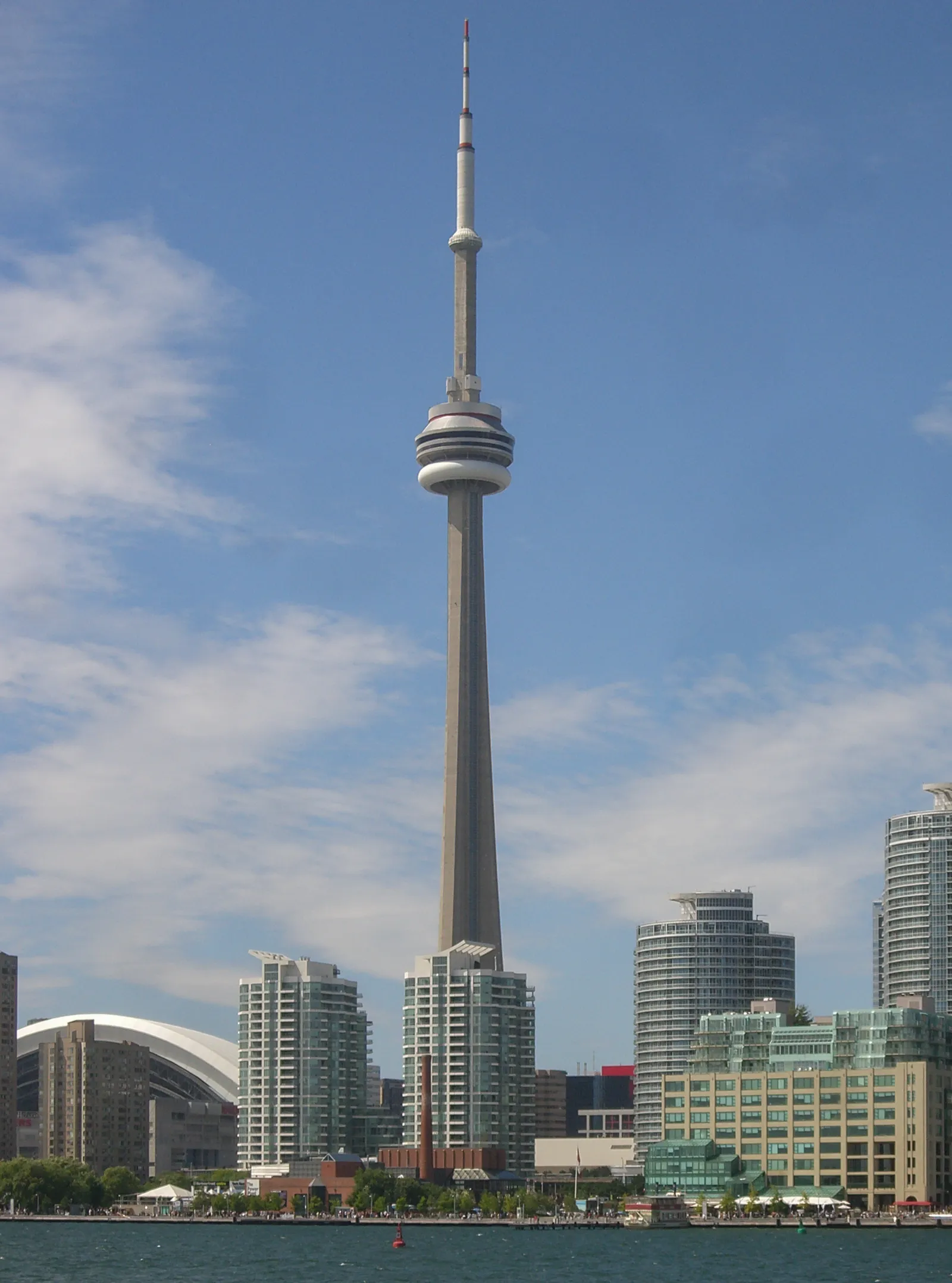The CN Tower rising above the Toronto waterfront, its round observation pod and antenna mast clear against a blue sky with the white roof of the Rogers Centre and harbourfront apartment blocks below