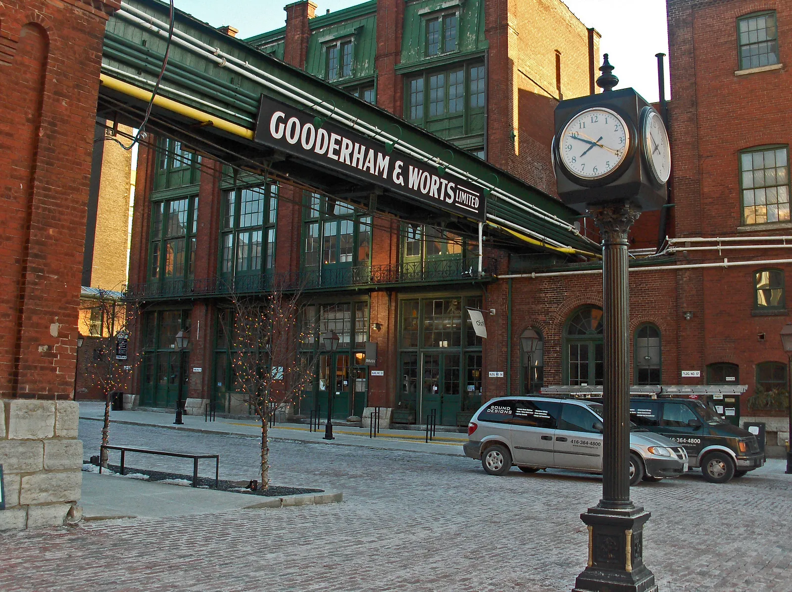 A brick-paved street in Toronto's Distillery District with a green cast-iron canopy carrying the Gooderham and Worts name over the entrance to a red-brick warehouse and a post clock in the foreground