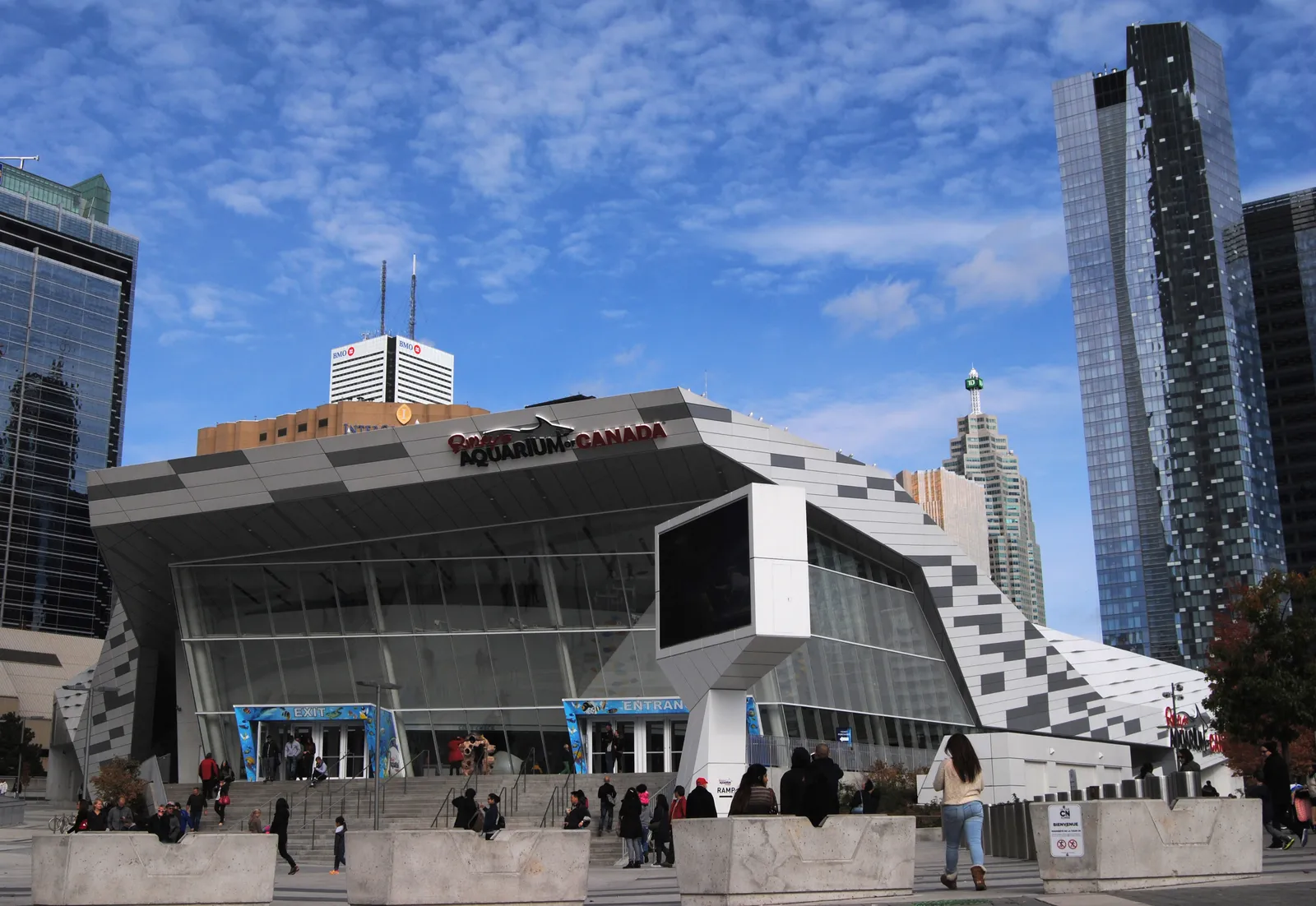 The angular white and glass facade of Ripley's Aquarium of Canada in Toronto with visitors on the steps outside and downtown office towers rising behind it