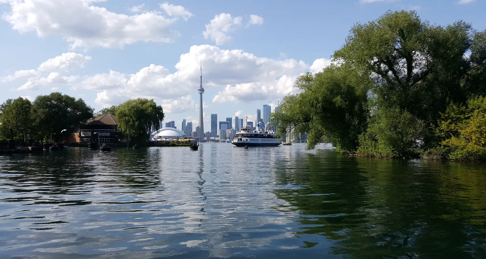 The Toronto skyline and CN Tower seen from a willow-lined lagoon on the Toronto Islands, with a passenger ferry crossing in front of the downtown towers