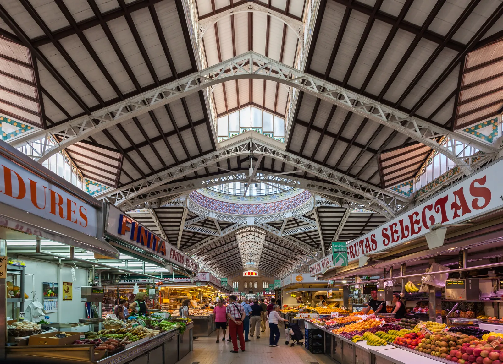 The main aisle of the Mercado Central in Valencia, fruit and vegetable stalls under a high iron roof with a tiled dome at the far end and shoppers walking between them