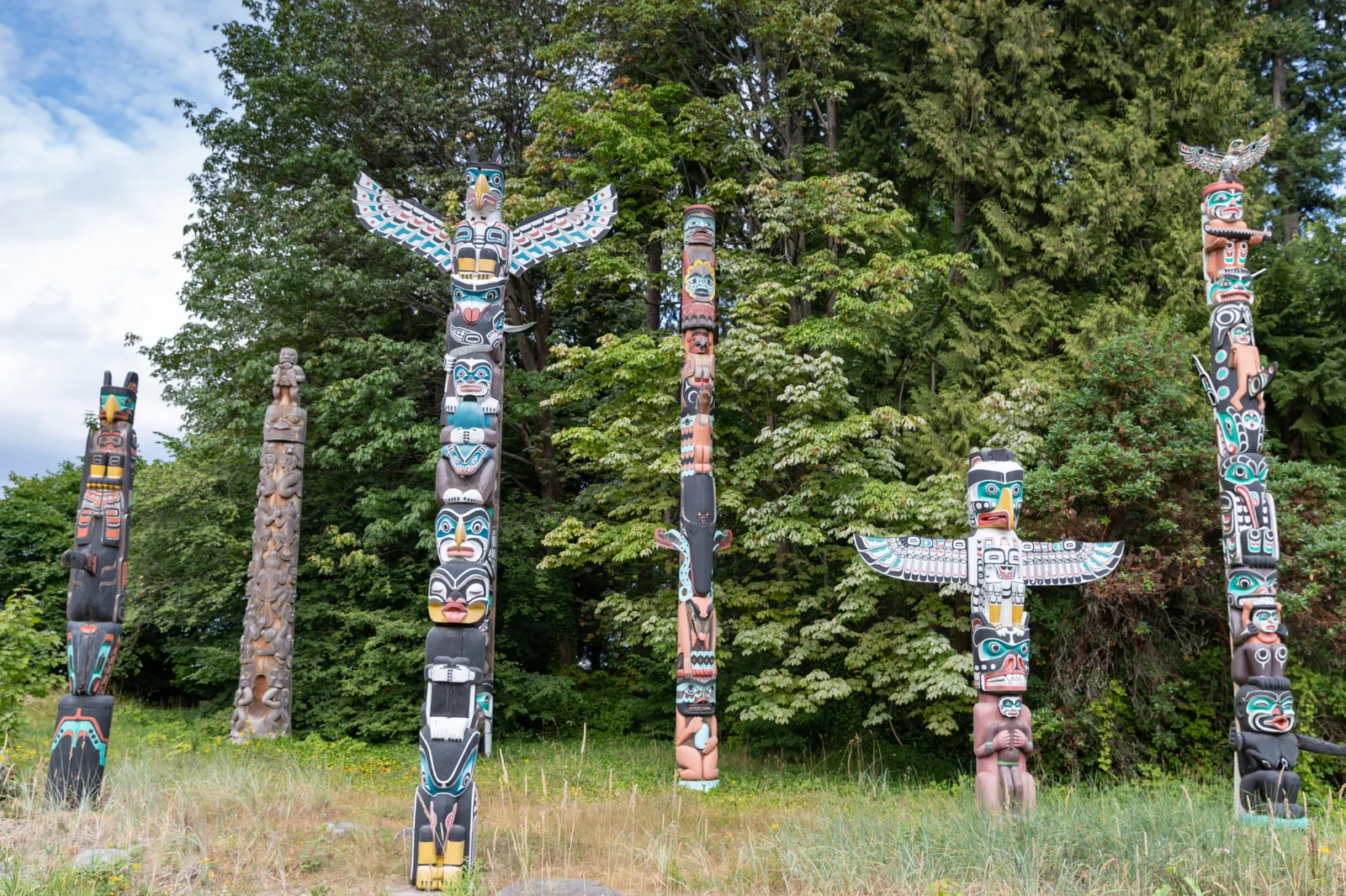 Six painted totem poles standing on grass at Brockton Point in Stanley Park, Vancouver, backed by a wall of dark evergreen forest