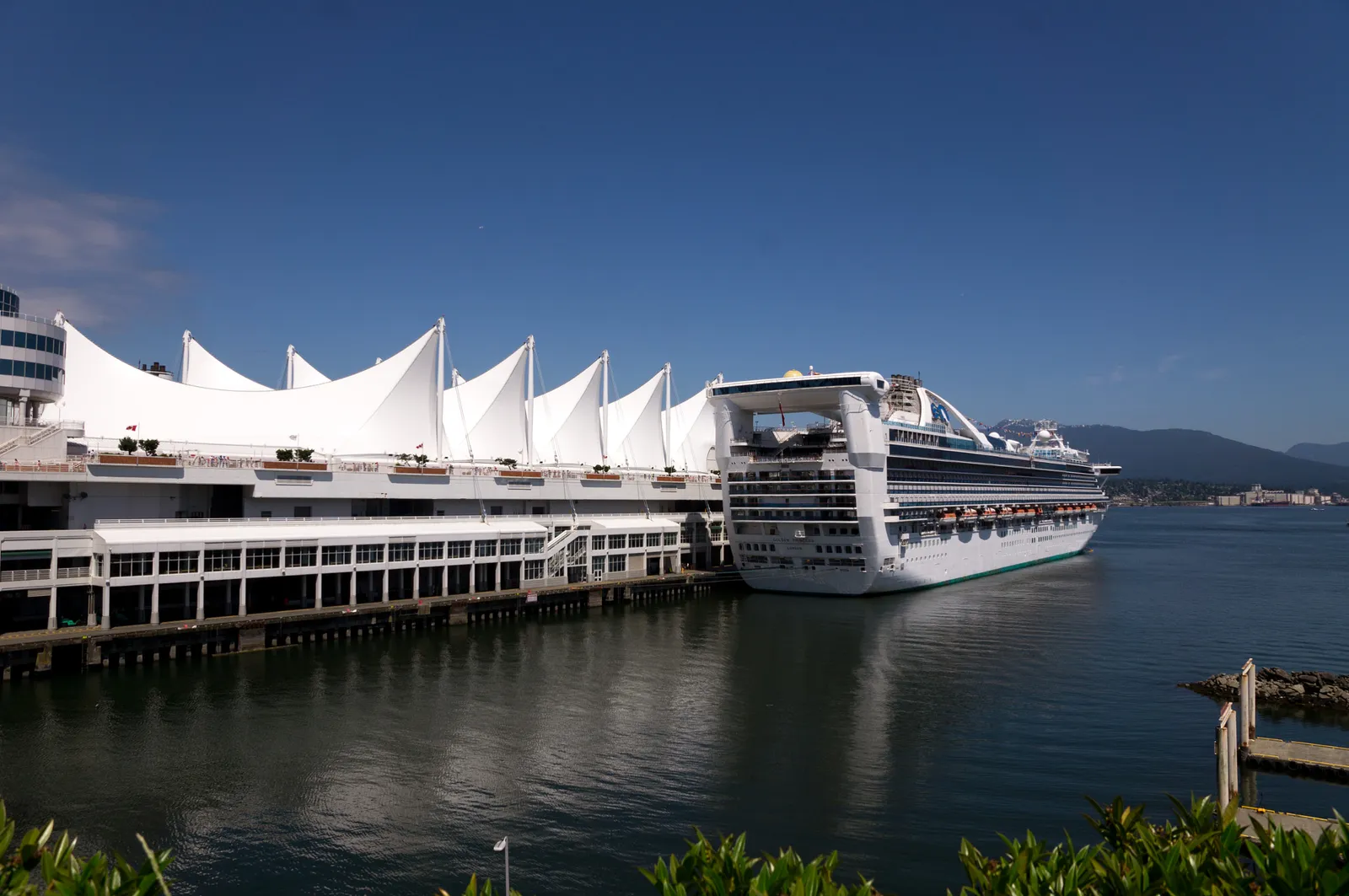 The five white fabric sails over the roof of Canada Place in Vancouver, with a cruise ship berthed alongside and the North Shore mountains across Burrard Inlet