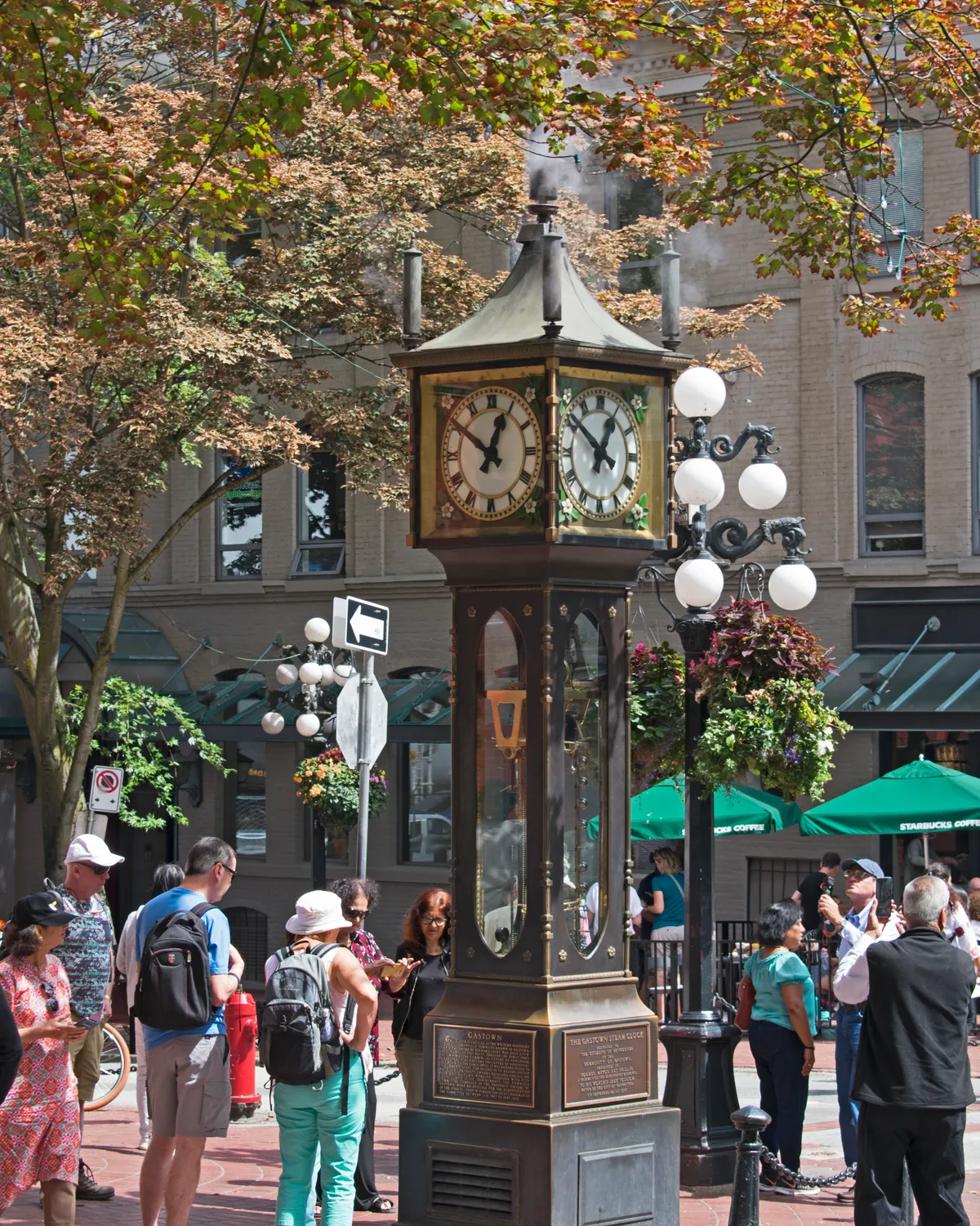 The brass and cast-iron Gastown steam clock on a Vancouver street corner, releasing a puff of steam from its roof vents while visitors stand around its base