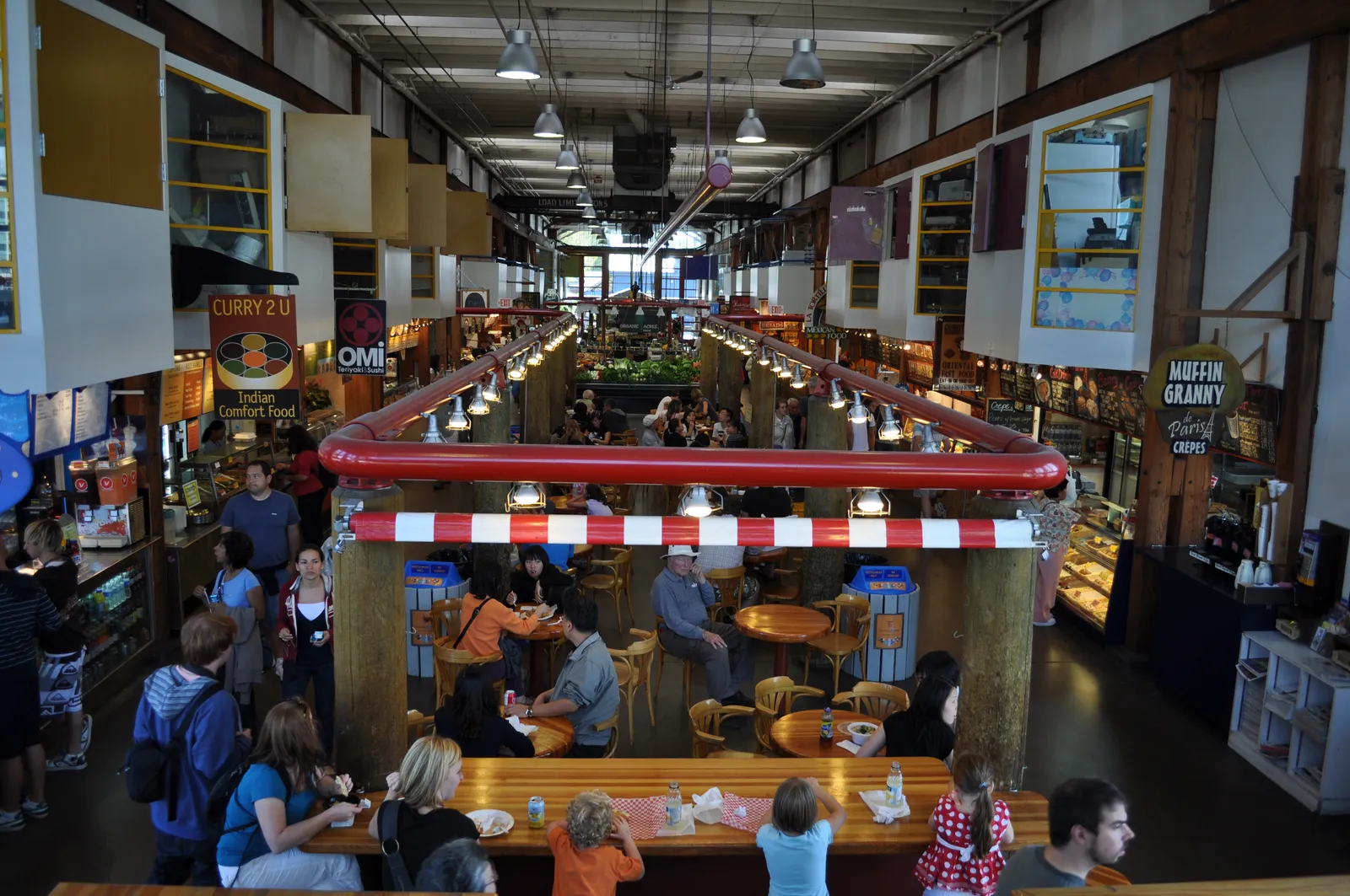 The interior of the Granville Island Public Market in Vancouver seen from a balcony, with food stalls under timber roof trusses and shoppers eating at long communal tables