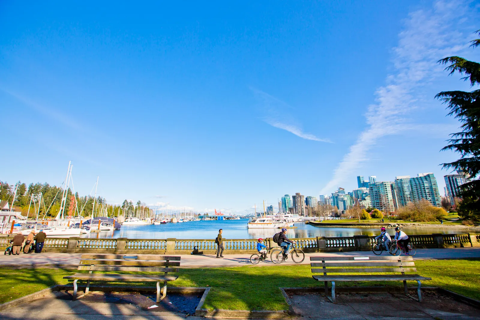 The paved Stanley Park seawall path in Vancouver on a clear day, with benches and cyclists in the foreground, moored boats behind and the downtown towers across Coal Harbour