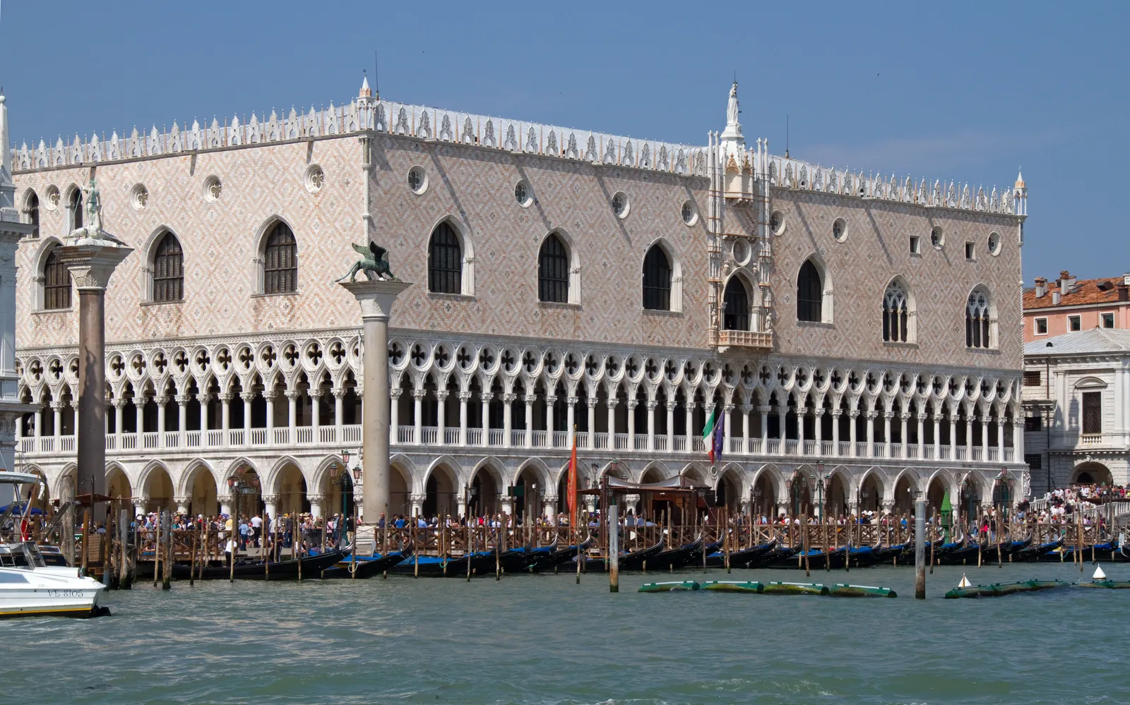 The waterfront front of the Doge's Palace in Venice seen from the lagoon, its two tiers of white stone arcades below a pink and white patterned upper wall, with mooring poles in the water in front