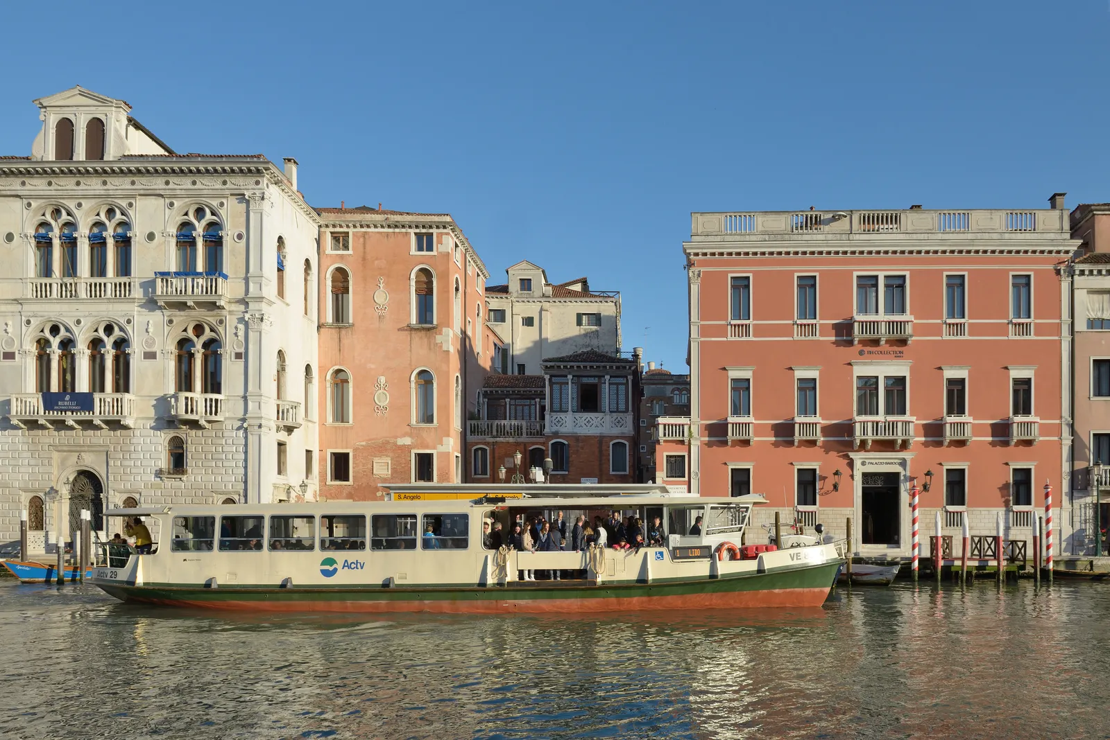 A public water bus full of standing passengers crossing the Grand Canal in Venice, with a row of pink and cream palace fronts along the far bank behind it