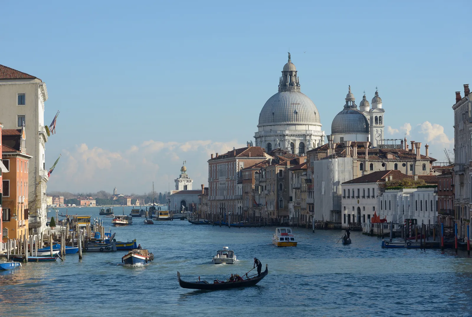 The Grand Canal in Venice seen from the crown of the Accademia bridge, palace fronts lining both banks and the grey dome of Santa Maria della Salute closing the view at the far end, with a gondola and water buses on the water