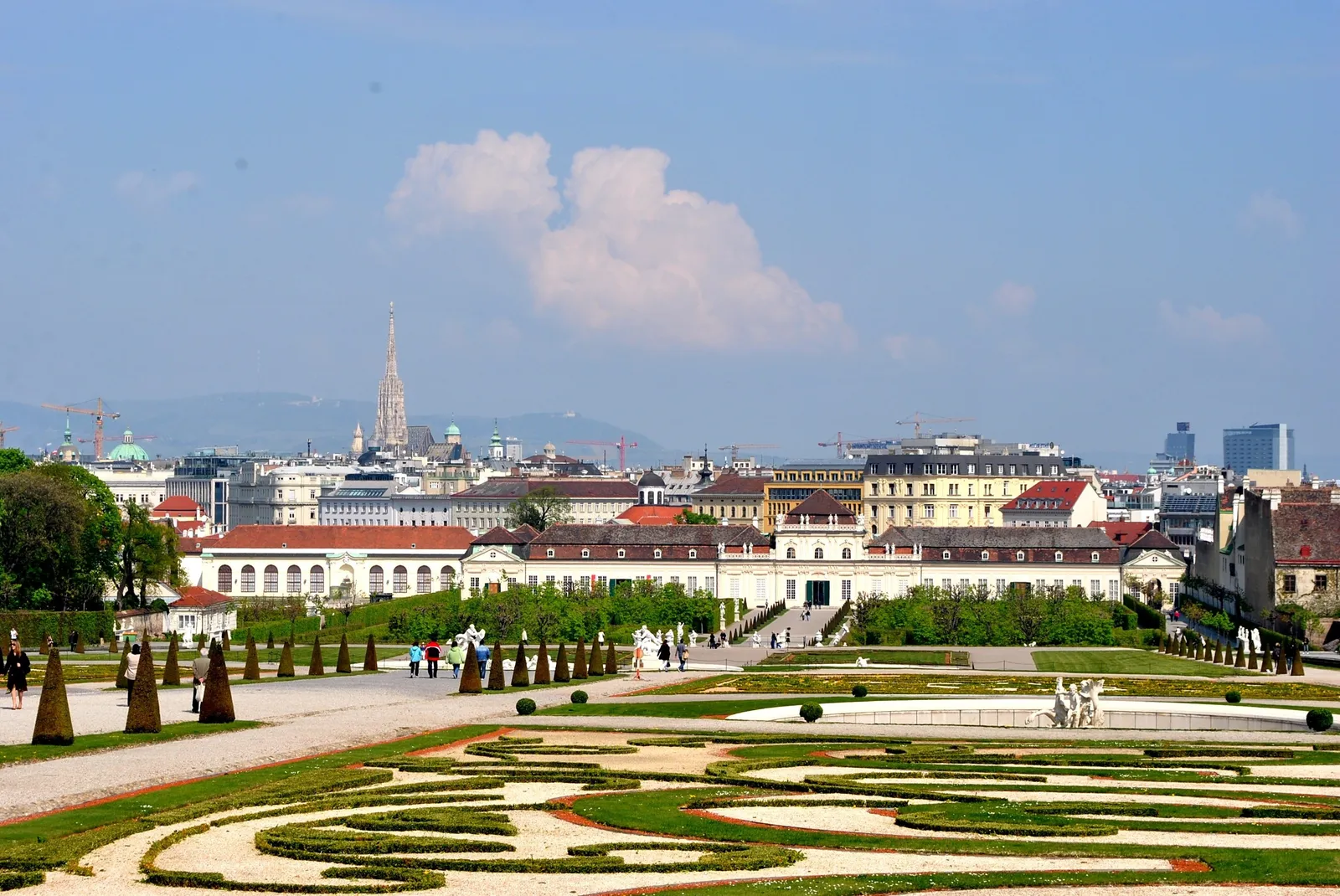 The view north from the Upper Belvedere terrace in Vienna, down over the garden's clipped hedge patterns and conical shrubs to the rooftops of the city, with the spire of St. Stephen's Cathedral on the skyline