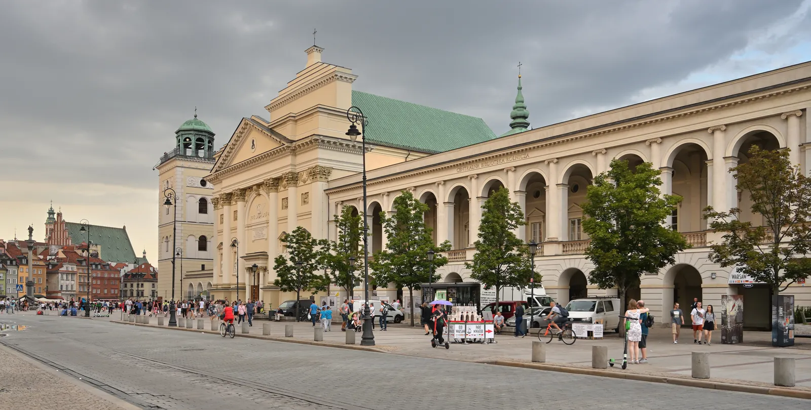 Krakowskie Przedmieście in Warsaw, the pale facade and green dome of St Anne's Church beside a long colonnaded wing, with people walking the wide traffic-free paving in front