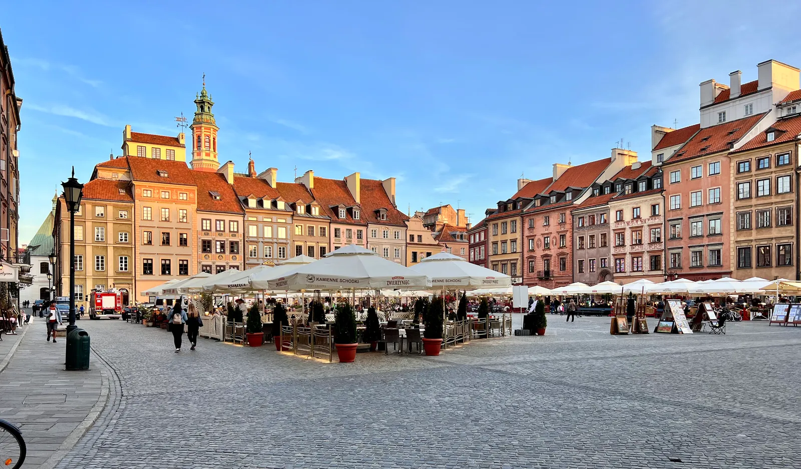 Warsaw's Old Town Market Square in late afternoon light, tall narrow townhouses painted ochre, pink and green around all four sides, café umbrellas in the middle and grey setts underfoot