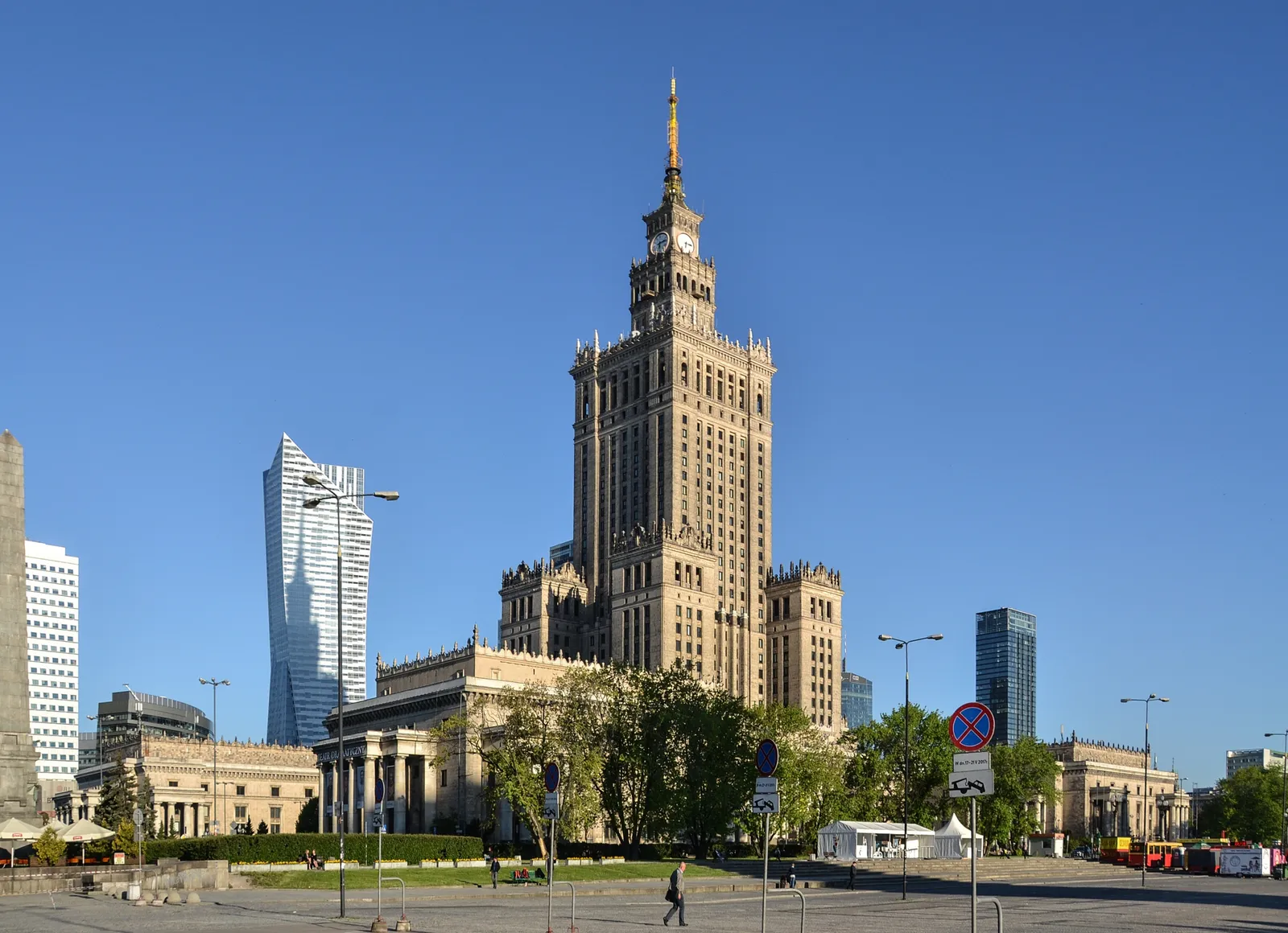 The Palace of Culture and Science in Warsaw, a tiered stone tower topped by a tall spire, with glass office towers standing alongside it against a clear sky