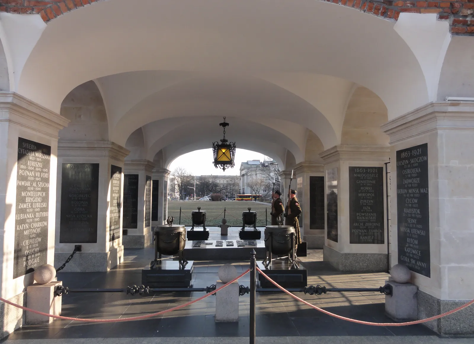 The Tomb of the Unknown Soldier in Warsaw under the surviving white arcade of the Saxon Palace, two eternal flames burning over the slab and battle names cut into black tablets on the pillars