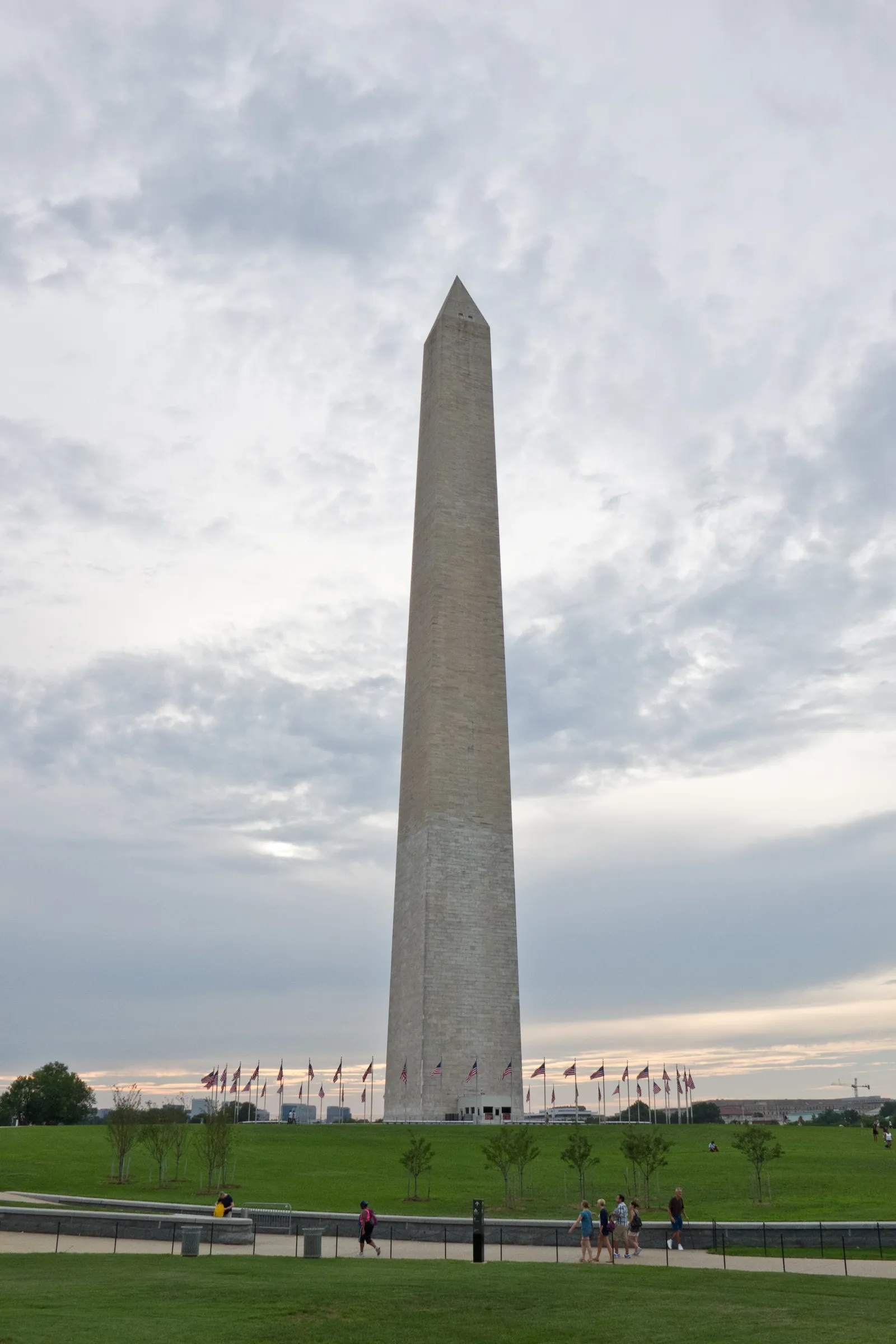 The Washington Monument seen from the lawn at its base, a plain stone obelisk rising to a pointed cap, ringed at the foot by a circle of American flags