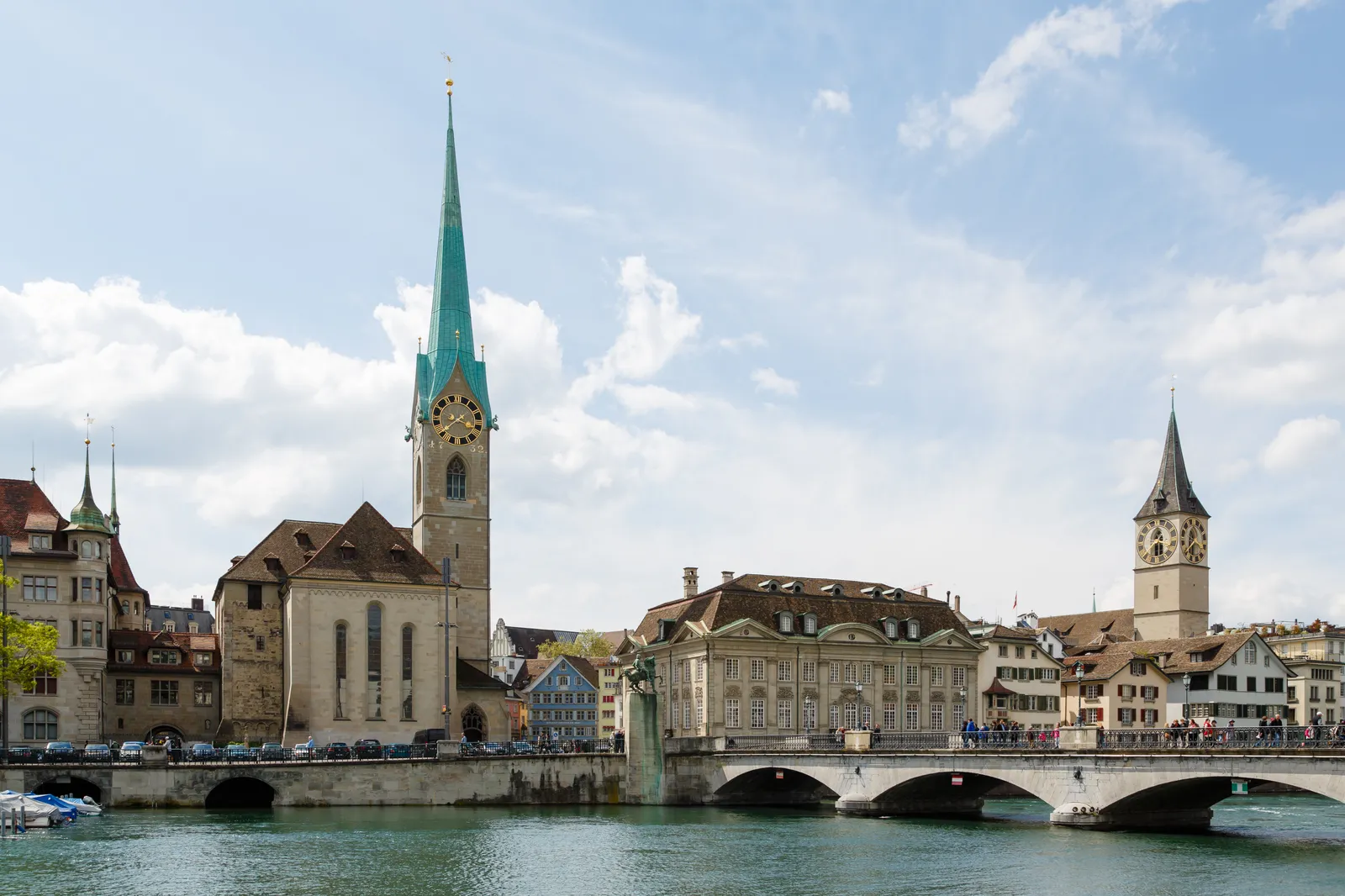The Fraumünster in Zurich from across the Limmat, its slim green copper spire and blue clock face above the stone church, with the Münsterbrücke and St. Peter's tower to the right