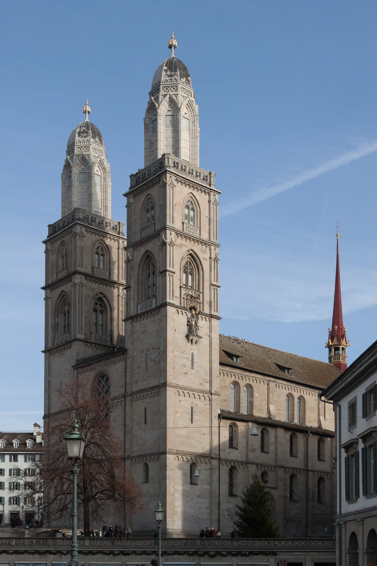 The Grossmünster in Zurich seen from the Münsterbrücke, its two square Romanesque towers rising to matching neo-Gothic tops against a clear blue sky, with the church's long stone flank running back to the right
