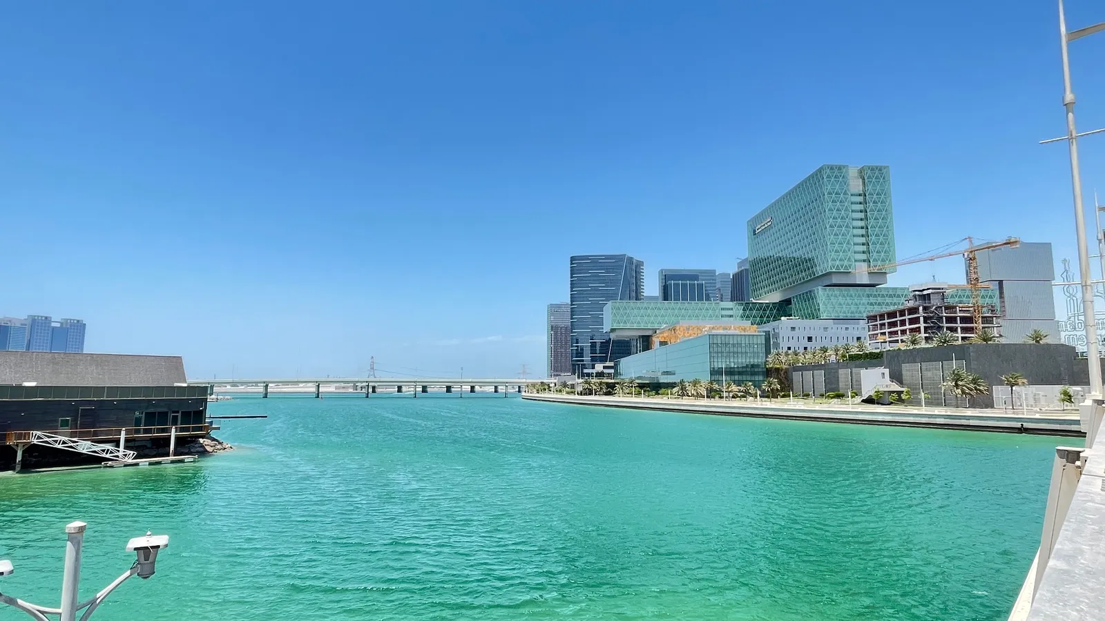 The glass towers and low waterfront buildings of Al Maryah Island in Abu Dhabi seen across a turquoise channel, with a road bridge running to the mainland on the left